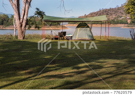 View of a camping tent with awning pitched on the dry ground near beautiful big trees with branches at the campsite lake shore with mountain range in background. View of a camping tent with awning pitched on the dry ground near beautiful big trees with branches at the campsite lake shore with mountain range in background. 77620579