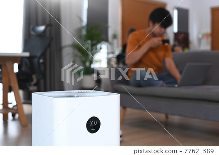 Air purifier on the floor in living room and young man sitting on sofa in background. 77621389