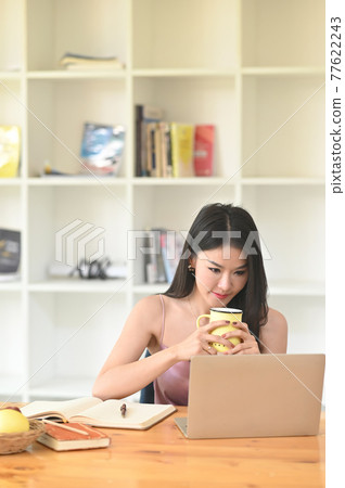 A woman is holding a coffee cup in her hands while sitting in front of a computer laptop. 77622243