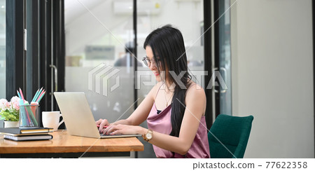 A beautiful woman is typing on a computer laptop while sitting at the wooden working desk. A beautiful woman is typing on a computer laptop while sitting at the wooden working desk. 77622358