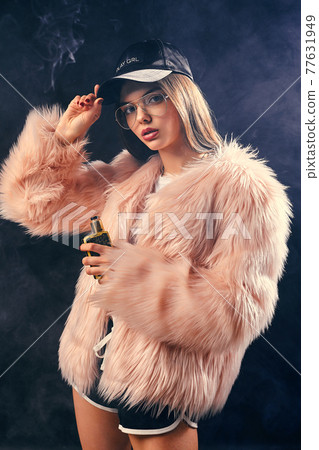 Sexy trendy rap woman in pink fur coat and cap smoke electronic cigarette. Dark Studio background shooting 77631949