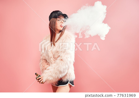 Sexy trendy rap woman in pink fur coat and cap smoke electronic cigarette. Pink Studio background shooting 77631969