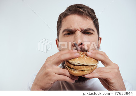 A man eating a hamburger on a light background in a white T-shirt cropped view close-up hunger fast food A man eating a hamburger on a light background in a white T-shirt cropped view close-up hunger fast food 77633944