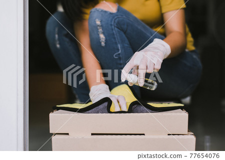 Woman spraying alcohol to paper box from delivery service during coronavirus pandemic situation at home 77654076