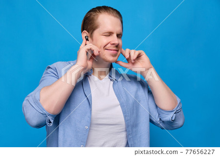 A man in casual clothes on a blue background with closed eyes presses the wireless headphones into his ears and sings along. A man in casual clothes on a blue background with closed eyes presses the wireless headphones into his ears and sings along. 77656227