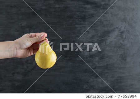 A person holding a yellow pear from the stem A person holding a yellow pear from the stem 77658994