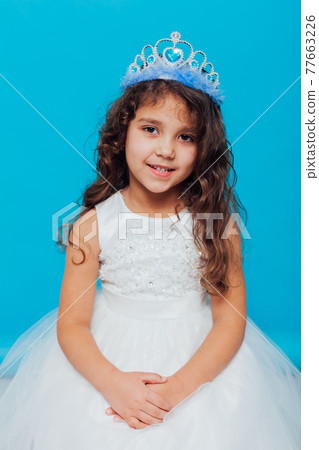 Beautiful little curly girl with crown tiara in white dress on a blue background background Beautiful little curly girl with crown tiara in white dress on a blue background background 77663226