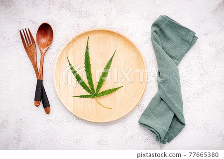 Food conceptual image of a cannabis leaf  with spoon and fork on white concrete background. 77665380