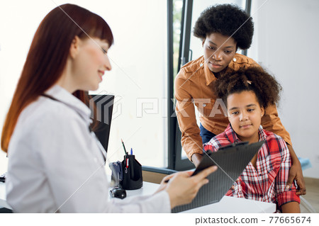 Female doctor pediatrician showing medical report or medical test results to young African mother and her tenn daughter on a clipboard in modern clinic. Children doctor talking to patient. 77665674