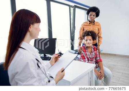 View from the doctor's shoulder of African-American mother and cute teen girl listening to the pediatrician. recommendations. Female Caucasian doctor fill in paper form and takes anamnesis. 77665675