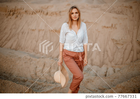 Young female exploring sandy beach with hat. Hipster girl, young traveler in nature desert. A woman walks in the desert to meet. Woman walks. 77667331