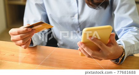 Cropped image of a woman is holding a credit card and smartphone in hands while sitting in the living room. Cropped image of a woman is holding a credit card and smartphone in hands while sitting in the living room. 77681688