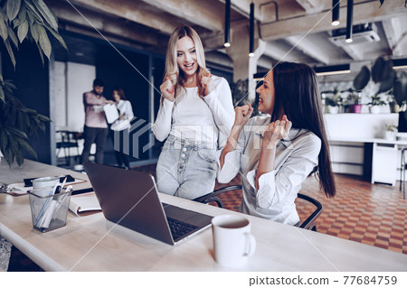 Two young happy businesswomen celebrating project success in office Two young happy businesswomen celebrating project success in office 77684759