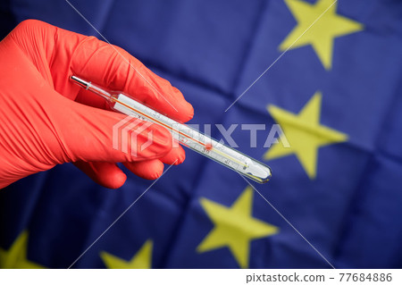 A hand in red gloves holds a thermometer with a high temperature on the background of the European Union flag. 77684886