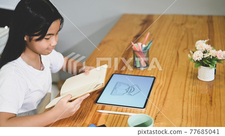 A schoolgirl is doing an E-Learning with a computer tablet at the wooden working desk. A schoolgirl is doing an E-Learning with a computer tablet at the wooden working desk. 77685041