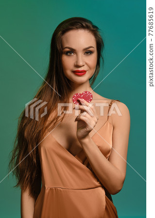 Brunette female in silk beige dress. She smiling, showing two red chips, posing against green background. Poker, casino. Close-up 77685619