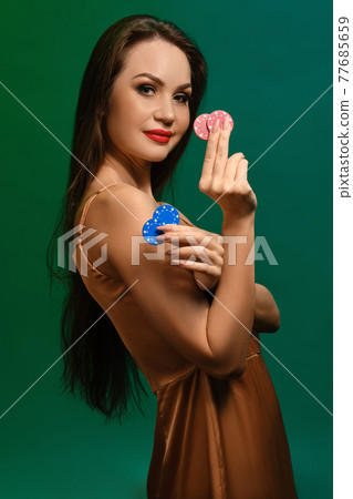 Brunette girl in silk beige dress. Smiling, showing four colorful chips, posing sideways on green background. Poker, casino. Close-up 77685659