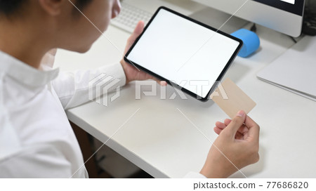 A young smart man is shopping online with a computer tablet at the white working desk. A young smart man is shopping online with a computer tablet at the white working desk. 77686820