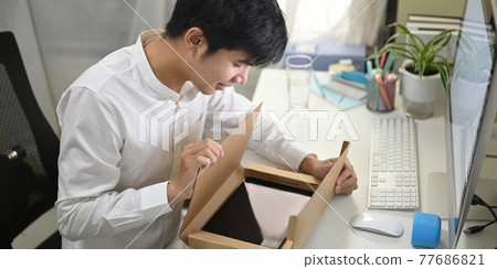 A smart man is unboxing a cardboard box from shopping online at the white working desk. A smart man is unboxing a cardboard box from shopping online at the white working desk. 77686821
