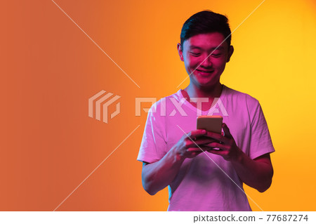 Portrait of Asian young man with smartphone isolated over orange yellow background in neon light. Portrait of Asian young man with smartphone isolated over orange yellow background in neon light. 77687274