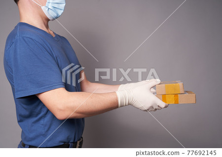Hands of a man in mask and gloves with two parcels on a gray background 77692145