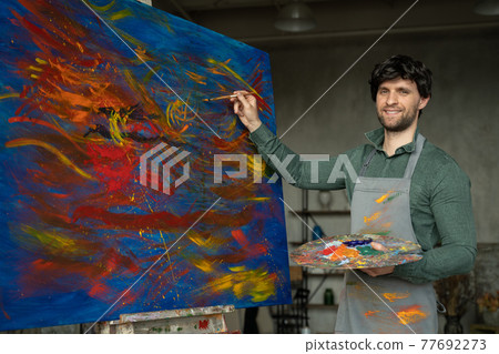 Portrait of male artist working on painting in studio. Male artist makes strokes with yellow paint 77692273