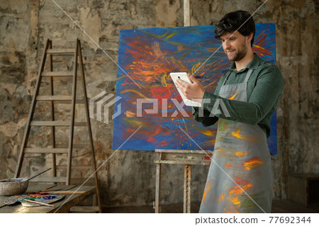 Male artist is standing in his studio and using a tablet, working on a project. Workshop with oil painting 77692344