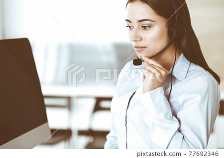 Brunette female customer service representative in a headset is consulting clients online. Call center and business people concept Brunette female customer service representative in a headset is consulting clients online. Call center and business people concept 77692346