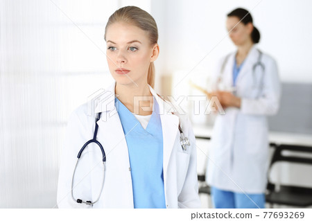 Woman - doctor standing in sunny clinic with colleague at the background. Woman - doctor standing in sunny clinic with colleague at the background. 77693269