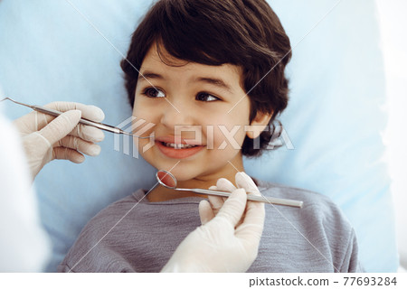 Cute arab boy sitting at dental chair with open mouth during oral checking up with doctor. Visiting dentist office. Stomatology concept Cute arab boy sitting at dental chair with open mouth during oral checking up with doctor. Visiting dentist office. Stomatology concept 77693284