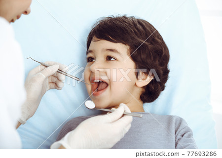 Cute arab boy sitting at dental chair with open mouth during oral checking up with doctor. Visiting dentist office. Stomatology concept 77693286