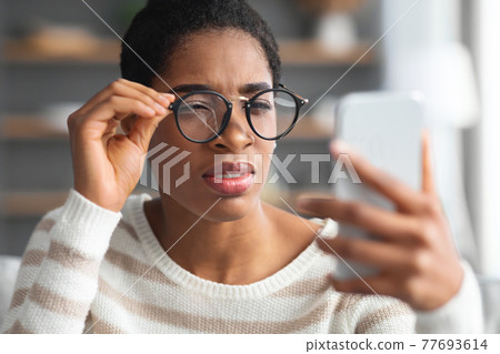 Astigmatism. Young Black Female In Eyeglasses Trying To Read Message On Smartphone Astigmatism. Young Black Female In Eyeglasses Trying To Read Message On Smartphone 77693614