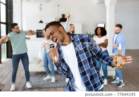 African American guy with beer bottle singing karaoke on home party with friends, singing songs into microphone 77694195