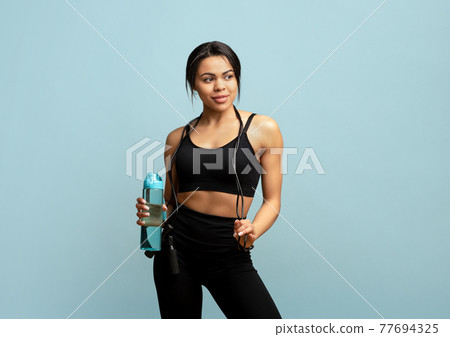 Fitness concept. Fit african american woman with jump rope and bottle of water posing over blue background Fitness concept. Fit african american woman with jump rope and bottle of water posing over blue background 77694325