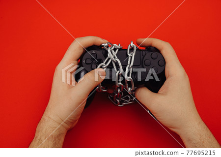 Game addiction concept. Hands of a young man holds a game controller is tied by chain Game addiction concept. Hands of a young man holds a game controller is tied by chain 77695215