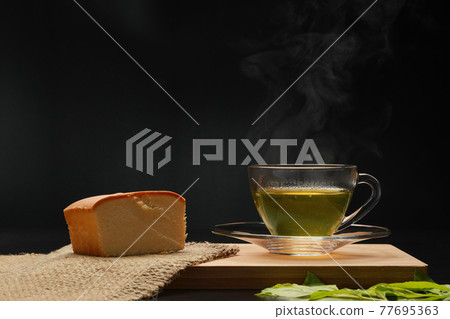 The hot green tea in the glass cup with smoke and butter cake on the wooden board as black background. Healthy drink concept. Dark tone. 77695363