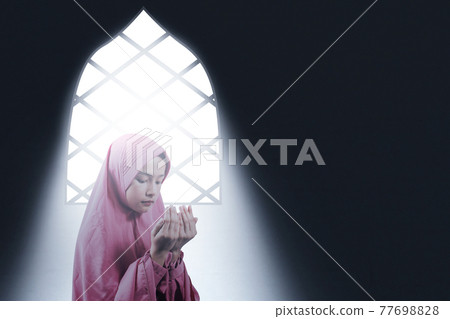 Asian Muslim woman in a veil sitting while raised hands and praying inside the room Asian Muslim woman in a veil sitting while raised hands and praying inside the room 77698828