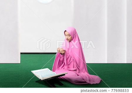 Asian Muslim woman in a veil sitting while raised hands and praying 77698833