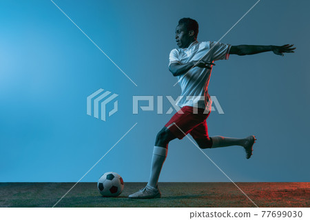 Young African soccer football player posing isolated on blue background in neon light. Rear view Young African soccer football player posing isolated on blue background in neon light. Rear view 77699030