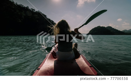 Silhouette of woman tourist raising hands while sailing in canoe along sea bay water to distant hilly island backside view. 77700338