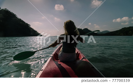 Silhouette of woman tourist raising hands while sailing in canoe along sea bay water to distant hilly island backside view. Silhouette of woman tourist raising hands while sailing in canoe along sea bay water to distant hilly island backside view. 77700339
