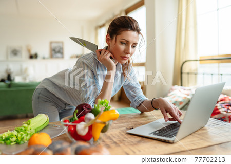 A young woman reads a recipe online on her laptop. Cooking lunch with fresh vegetables. A young woman reads a recipe online on her laptop. Cooking lunch with fresh vegetables. 77702213