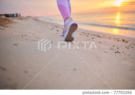 Closeup of runner legs in pink sports shoes performing jog on a sandy beach during sunrise in the early morning Closeup of runner legs in pink sports shoes performing jog on a sandy beach during sunrise in the early morning 77703266
