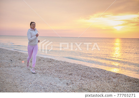 Full length attractive athletic woman doing morning jog along the coastline of the sea against the backdrop of a beautiful sunrise 77703271