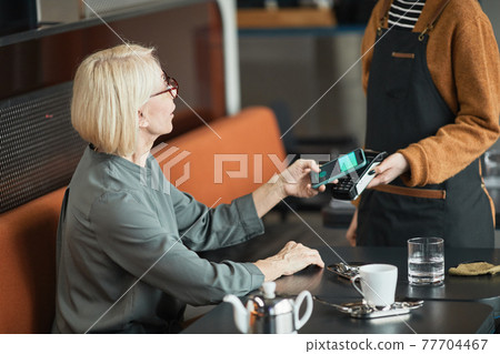Senior woman putting smartphone to payment terminal Senior woman putting smartphone to payment terminal 77704467