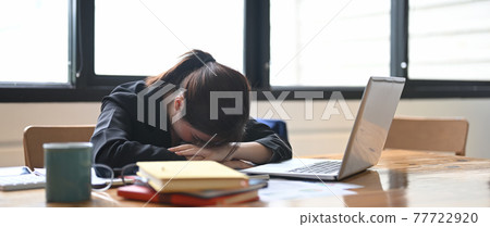 A stressed businesswoman is sitting at the office desk surrounded by a computer laptop and various equipment. 77722920