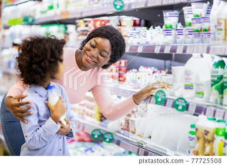 Positive young black mom teaching her daughter how to choose dairy products at grocery store Positive young black mom teaching her daughter how to choose dairy products at grocery store 77725508