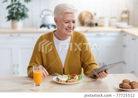 Positive senior woman having lunch and using digital tablet 77725512