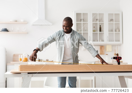 Assembling furniture. Happy african american man measuring table with roulette, standing and installing desk in kitchen 77726423