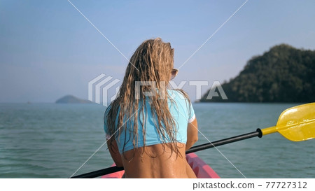 Long curly haired blonde woman in swimsuit rows plastic kayak along azure ocean water at exotic resort close backside view. 77727312